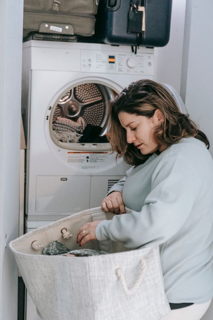 Side view of serious female with bag of dirty clothes loading modern washing machine while sitting in light washroom at home